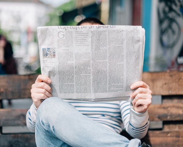 Eine Person sitzt auf einer Holzbank und wird dabei von ihrer Zeitung verdeckt